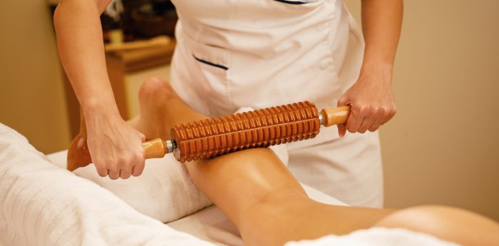 Massage minceur par madérothérapie : soin naturel pour sculpter le corps Praticienne réalisant un massage de madérothérapie sur les jambes d’une femme à l’aide d’un rouleau en bois cranté.